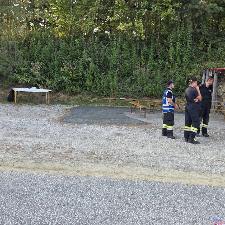 Ein Mann in einer blauen Weste bedient eine Drohne, während er in einem Kiesplatz steht. Hinter ihm stehen vier Personen in schwarzen Uniformen, möglicherweise Feuerwehrleute, in der Nähe eines roten Fahrzeugs.