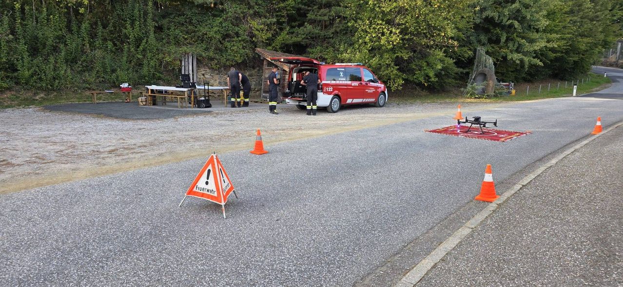 Ein roter Van mit offener Heckklappe steht am Straßenrand. Ein Drohnen liegt auf dem Boden in der Nähe, und es gibt orangefarbene Verkehrskegel und ein Warnschild auf der Straße.