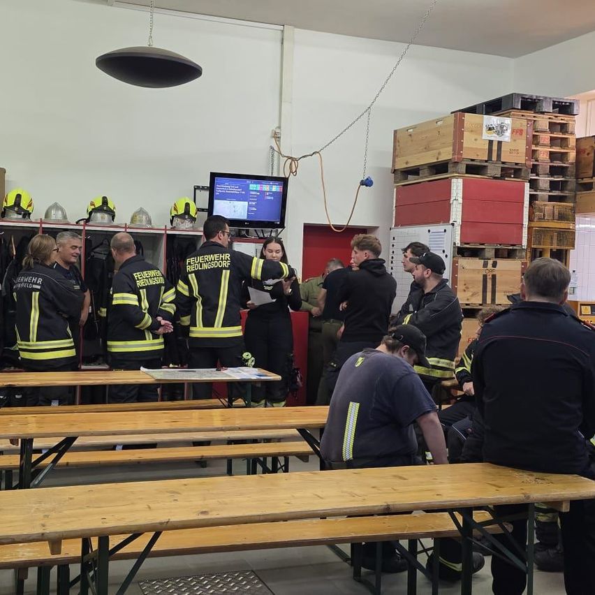 Eine Gruppe von Feuerwehrleuten versammelt sich in einem Raum mit Holzbänken, Helmen und Uniformen.