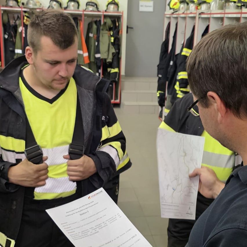 Drei Männer sind in einem Raum mit Feuerwehrausrüstung. Der Mann in der Mitte trägt eine reflektierende Sicherheitsjacke und hält ein Papier.