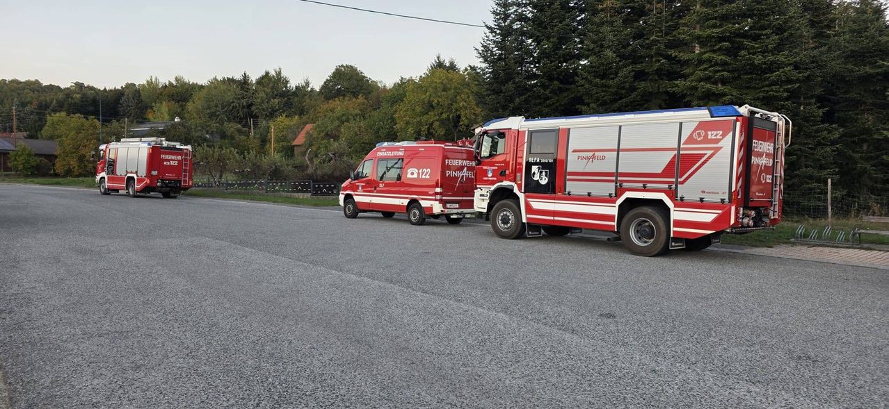 Drei rote Feuerwehrfahrzeuge sind am Straßenrand geparkt, mit Bäumen und einem Zaun im Hintergrund.
