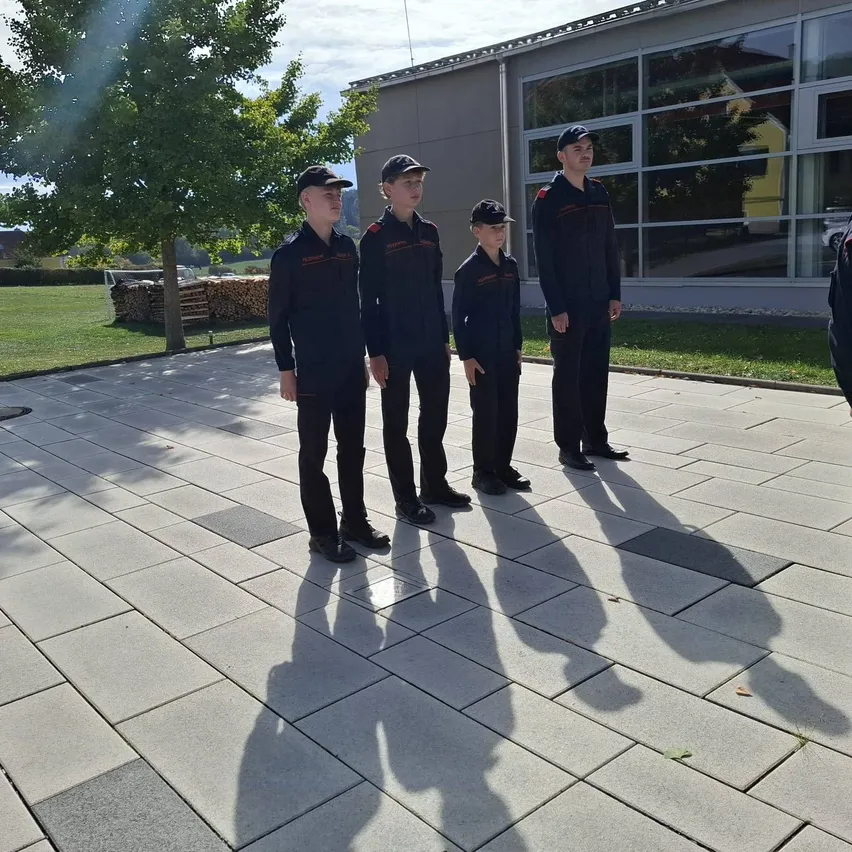 Fünf Personen in schwarzen Uniformen stehen in einer Reihe auf einem gefliesten Boden, mit einem Gebäude und einem Baum im Hintergrund.