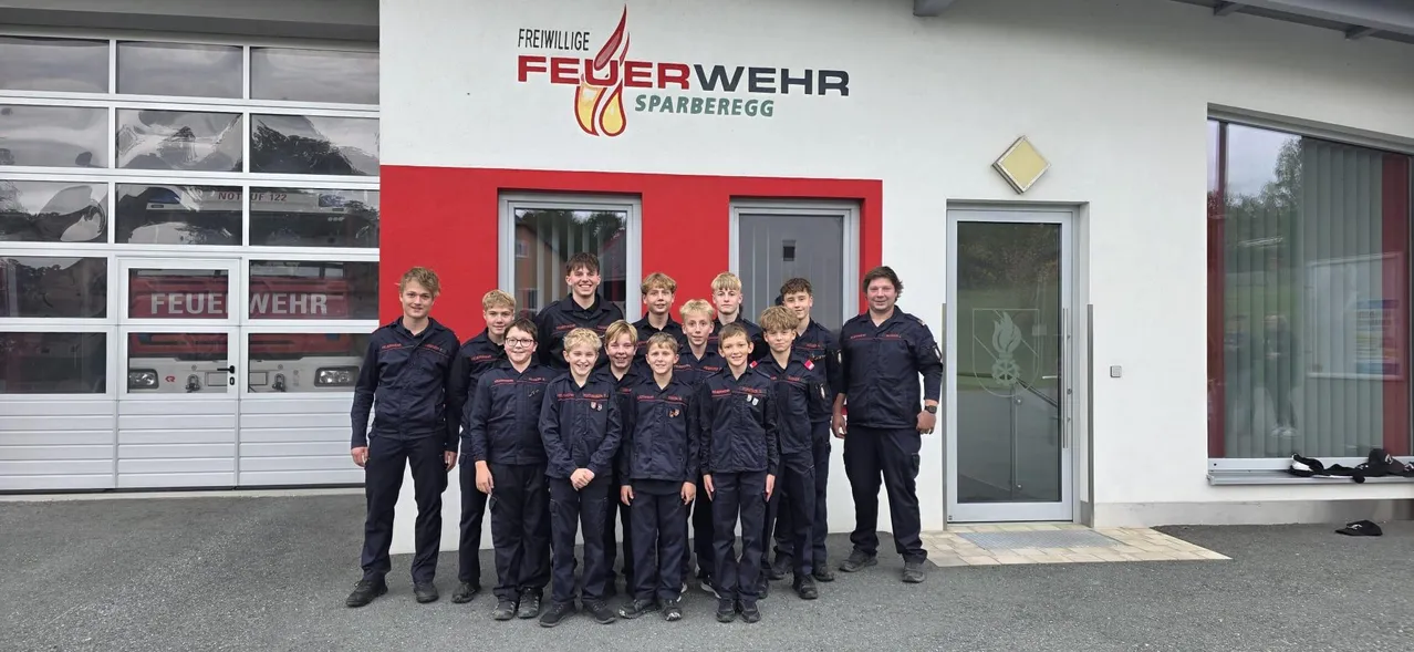 Eine Gruppe junger Jungen und Männer in passenden Uniformen posiert für ein Foto vor einem Feuerwehrhaus.