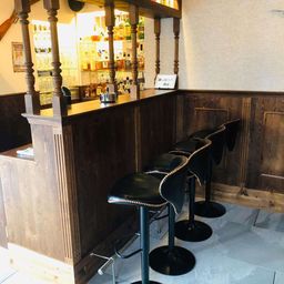 Eine hölzerne Bartheke mit vier schwarzen Leder-Barhockern davor. Regale mit Flaschen und Gläsern hinter der Theke. Holzwände und gefliester Boden.
