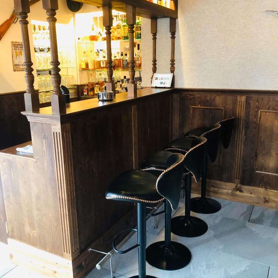 Eine hölzerne Bartheke mit vier schwarzen Leder-Barhockern davor. Regale mit Flaschen und Gläsern hinter der Theke. Holzwände und gefliester Boden.