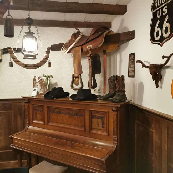 Ein rustikaler Raum mit einem Holzklavier, Cowboyhüten, Stiefeln und Sättel an der Wand. Eine Laterne und dekorative Gegenstände sind ebenfalls vorhanden.