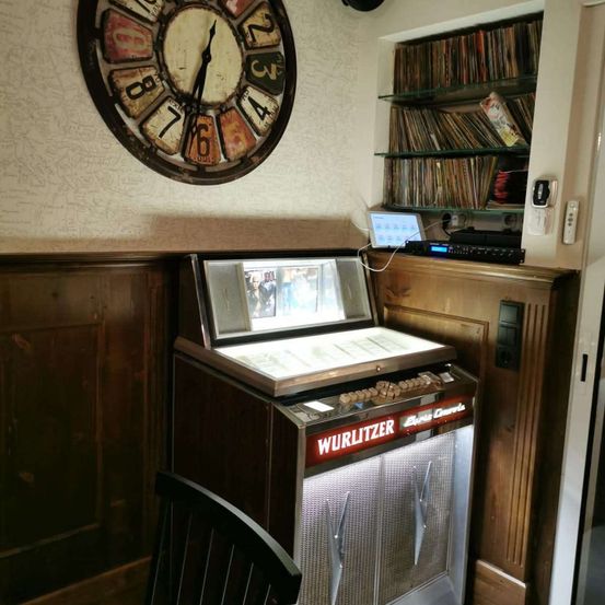 Ein Raum mit einem Wurlitzer-Jukebox, einer Uhr und einem Regal mit Schallplatten. Die Uhr zeigt 10:10 Uhr an.