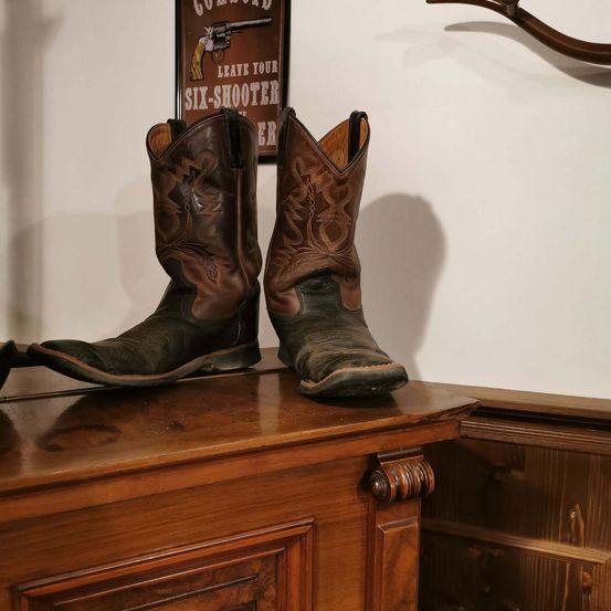 Zwei Cowboystiefel stehen auf einer Holzoberfläche. An der Wand dahinter befinden sich ein hölzerner Geweih und ein gerahmter Poster.