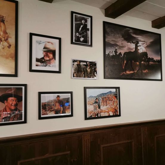 Eine Wand, geschmückt mit mehreren gerahmten Fotografien, darunter Porträts eines Cowboys und Szenen von Cowboys auf Pferden. Die Wand ist weiß gestrichen mit hölzerne Begrenzung.