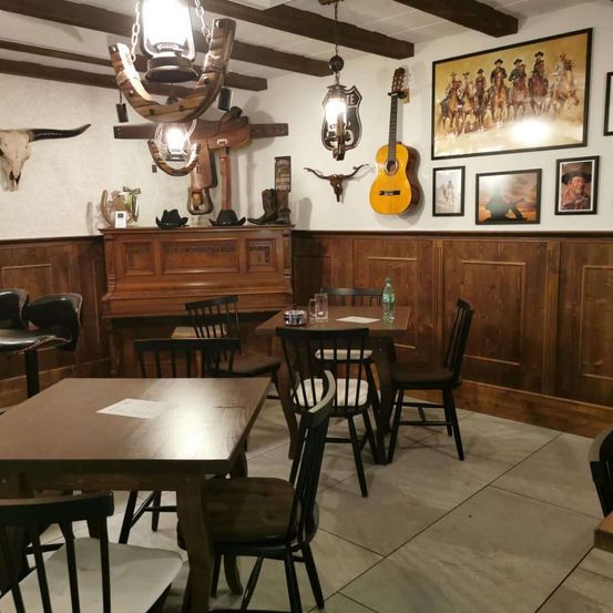 Ein leerer rustikaler Essbereich mit Holzwänden, Tischen und Stühlen. Dekoration umfasst Cowboy-Hüte, eine Gitarre und Western-Themen-Gemälde.