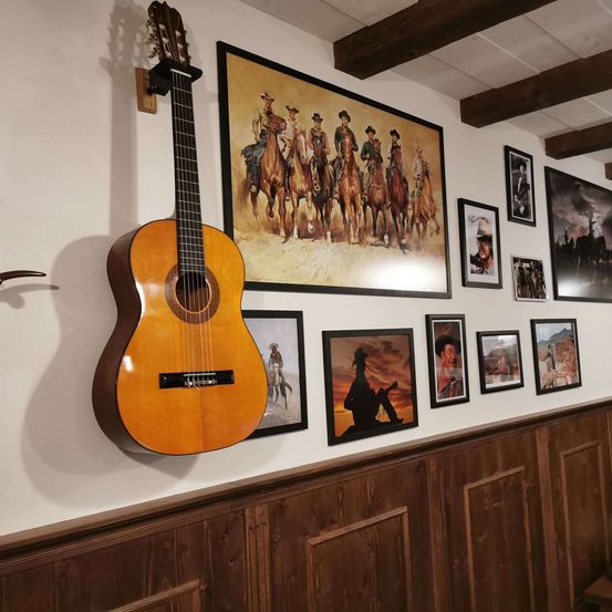 Eine Gitarre hängt an einer Wand, die mit Western-Themen-Kunstwerken geschmückt ist, darunter gerahmte Bilder von Cowboys auf Pferden. Die Wand hat Holzvertäfelung.