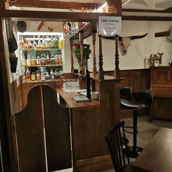 Das Innere einer Bar zeigt einen hölzernen Tresen mit Regalen, die mit Spirituosenflaschen bestückt sind. Ein Schild mit der Aufschrift 'CWRC Clublokal' hängt über dem Tresen. Ein Bullenschädel und ein Blumenstrauß sind auf dem Tresen zu sehen. Zwei schwarze Barhocker stehen in der Nähe.