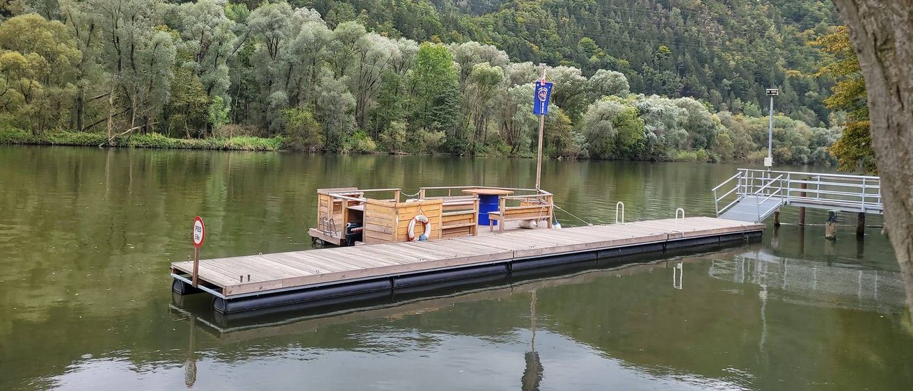 Eine Holzanlegestelle erstreckt sich in einen ruhigen See, umgeben von üppiger Grünfläche und Bergen. Sie hat einen Tisch, eine Bank und einen Rettungsring.