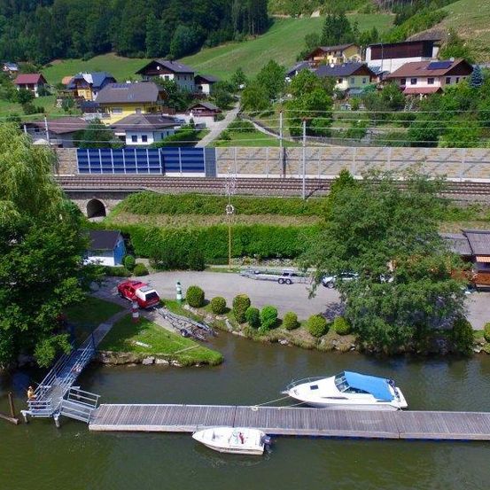Eine Luftaufnahme eines Flusses mit zwei angedockten Booten, einem Steg, und einem nahegelegenen Dorf mit Häusern und Bäumen. Ein rotes Auto ist am Ende des Stegs geparkt.