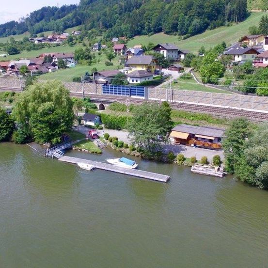 Eine Luftaufnahme eines Dorfes in der Nähe eines Flusses mit einem Steg, einem Boot, einem Haus und einer Bahnstrecke.
