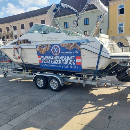 Ein weißes Boot auf einem Anhänger, das für die Marinekameradschaft Prinz Eugen Bruck wirbt, steht vor einem Gebäude mit vielen Fenstern.