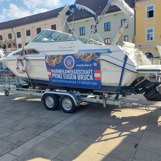 Ein weißes Boot auf einem Anhänger, das für die Marinekameradschaft Prinz Eugen Bruck wirbt, steht vor einem Gebäude mit vielen Fenstern.