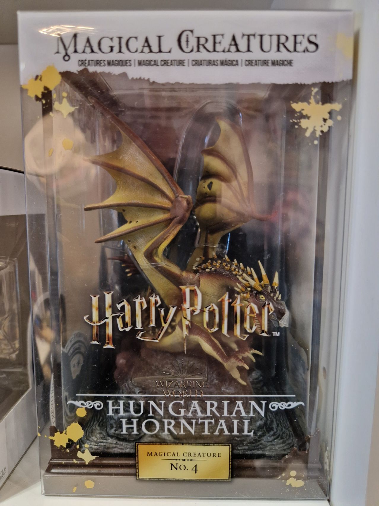 A plastic display case contains a Harry Potter Hungarian Horntail figurine. The figure is golden and has large wings and a spiked tail. The case has a yellow star design on the top.