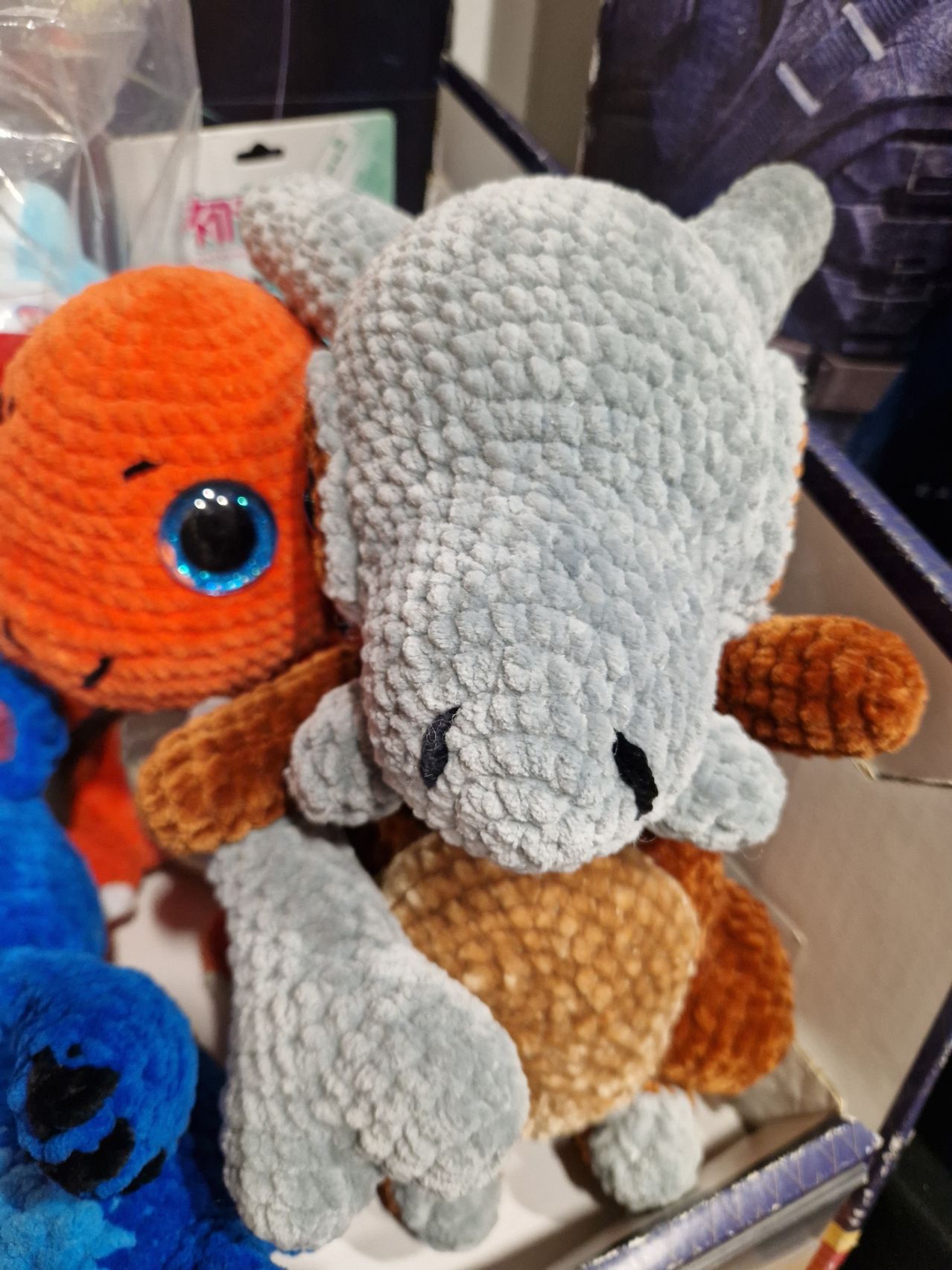 Ein graues Plüschtier mit großen blauen Augen und einem braunen Körper ruht auf einem orangefarbenen Plüschtier mit blauem Auge und kleinem Mund.