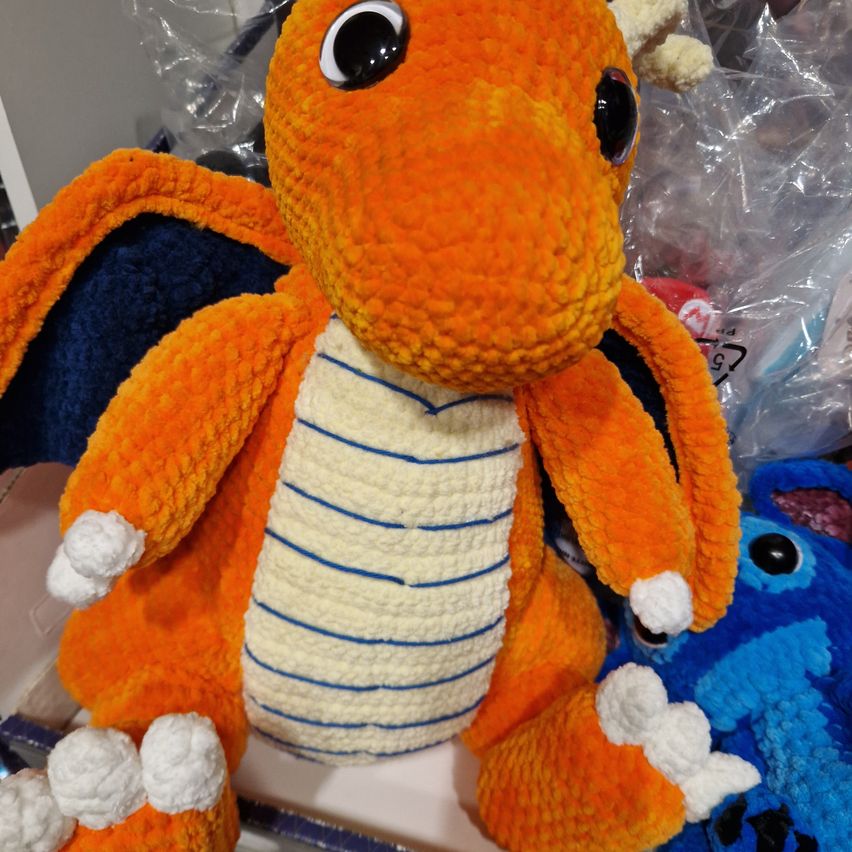 Ein gehäkeltes orangefarbenes Drachenplüschtier mit blauen Streifen auf dem Bauch, weißen Füßen und schwarzen Augen sitzt vor anderen Spielzeugen.
