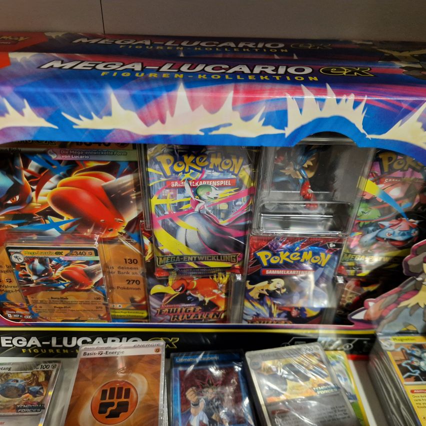 Eine Sammlung von Mega Lucario Pokemon Spielzeug und Karten im Display, einschließlich einer Figur, Sammelkarten und versiegelten Booster-Packungen. Die Produkte sind in Plastikverpackungen.