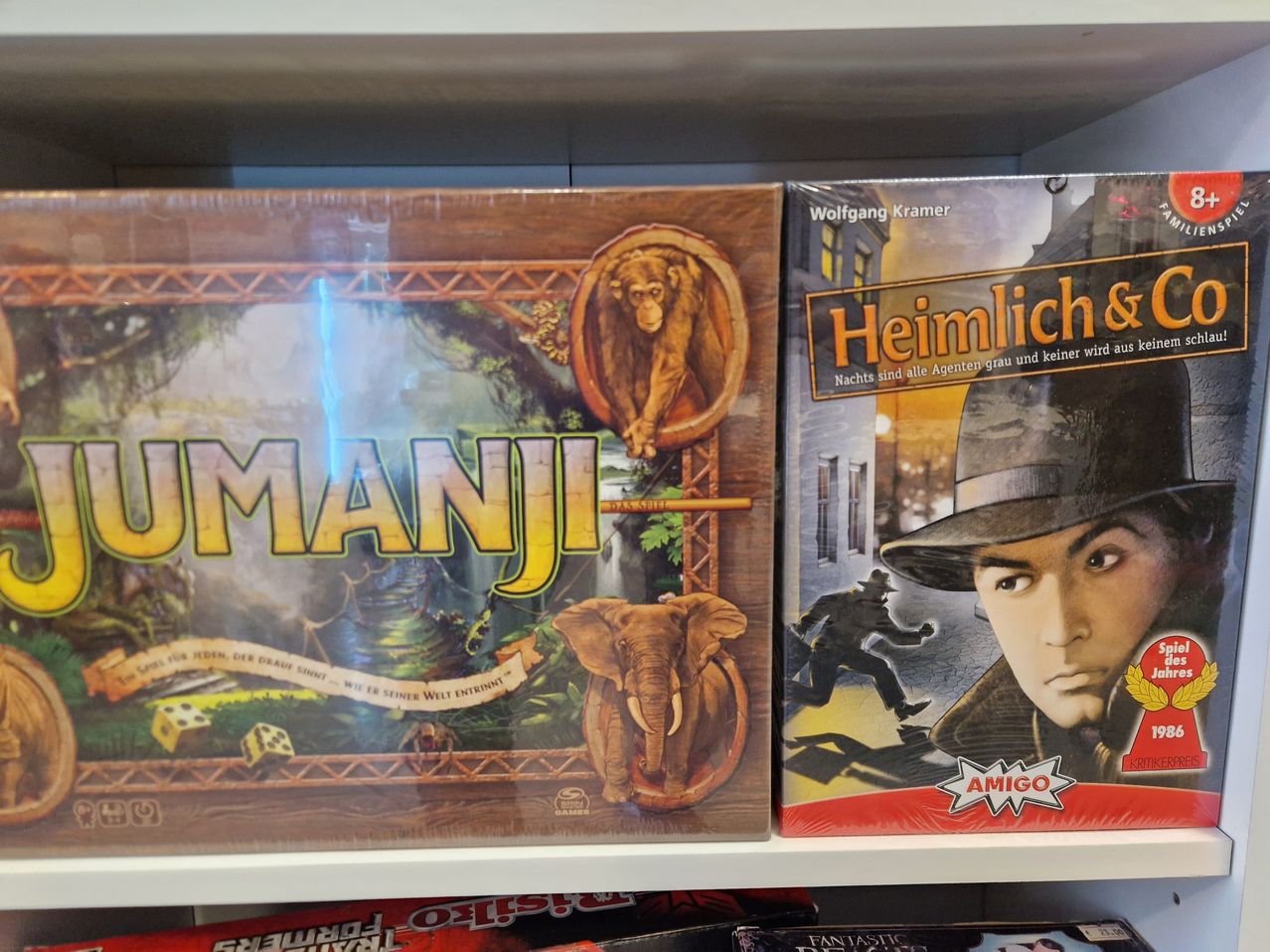 Zwei Brettspiele auf einem Regal, eines mit dem Titel 'Imanji' mit einem Elefanten und einem Affen, das andere 'Heimlich' mit einem Detektiv in einem Hut.