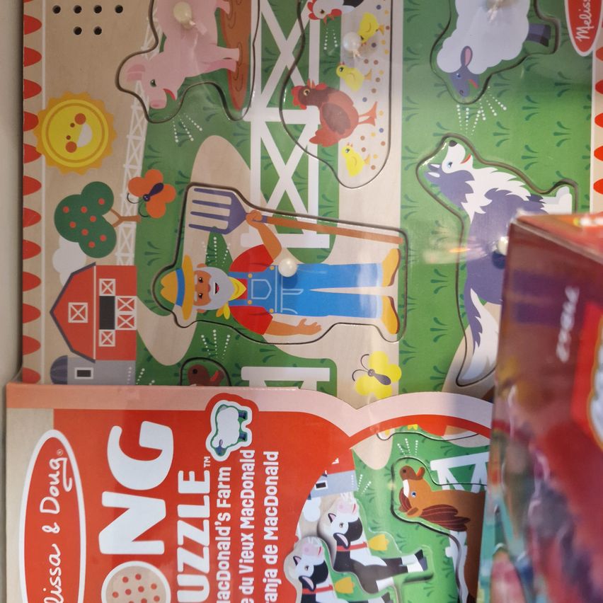 Ein pädagogisches Holzpuzzle zeigt eine Bauernhofszene mit Tieren und einem Bauern. Es zeigt einen Stall, eine Henne, eine Kuh, ein Pferd und eine Schaf. Unten steht der Name "Melissa und Doug".