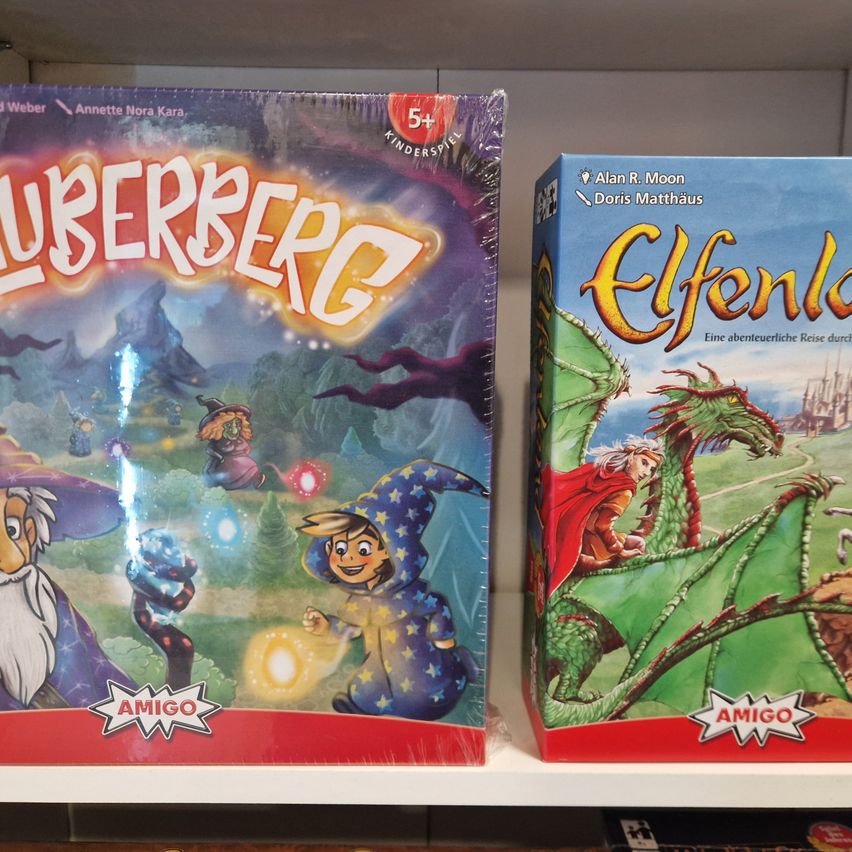 Zwei Brettspiele auf einem Regal ausgestellt. Das erste, 'Lubberbörg', zeigt eine farbenfrohe Fantasieszene. Das zweite, 'Elfenland', zeigt einen Drachen und einen Reiter auf blauem Hintergrund.