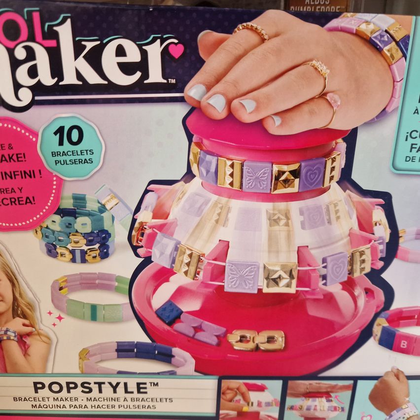 Ein buntes Armband-Herstellungsset mit mehreren gezeigten Armbändern, konzipiert für die Erstellung verschiedener Stile, einschließlich Perlen und Anhängern. Eine Hand mit Ringen hält die Oberseite der Maschine.