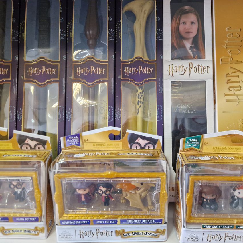 Mehrere Harry-Potter-Waren, darunter Actionfiguren und Zauberstäbe, sind in gelber und lila Verpackung ausgestellt. Die Figuren umfassen Hermine Granger, Harry Potter und andere.