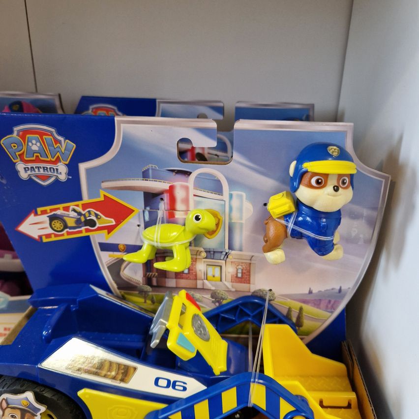 Ein blaues Spielzeugauto mit der Nummer 06 und einer Paw Patrol Hundefigur in blauer Uniform. Das Spielzeug ist in einer Schachtel mit einer grünen Schildkrötenfigur, die daran hängt.