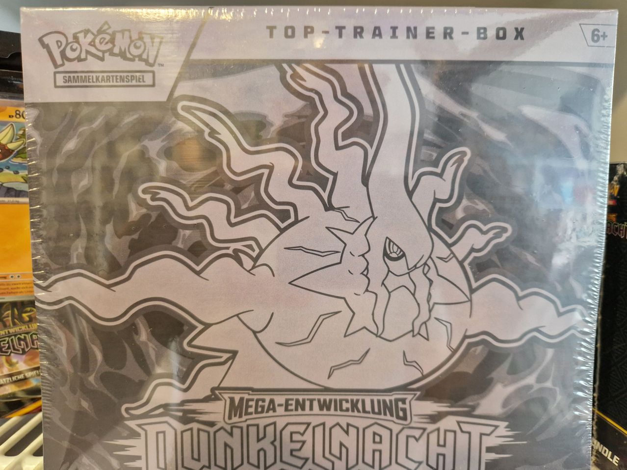 Ein Werbeplakat für eine Pokemon Top Trainer Box mit einer Schwarz-Weiß-Zeichnung eines Pokemons mit Blitzeffekten und dem Titel 'Mega-Entwicklung Dunkelnacht'.