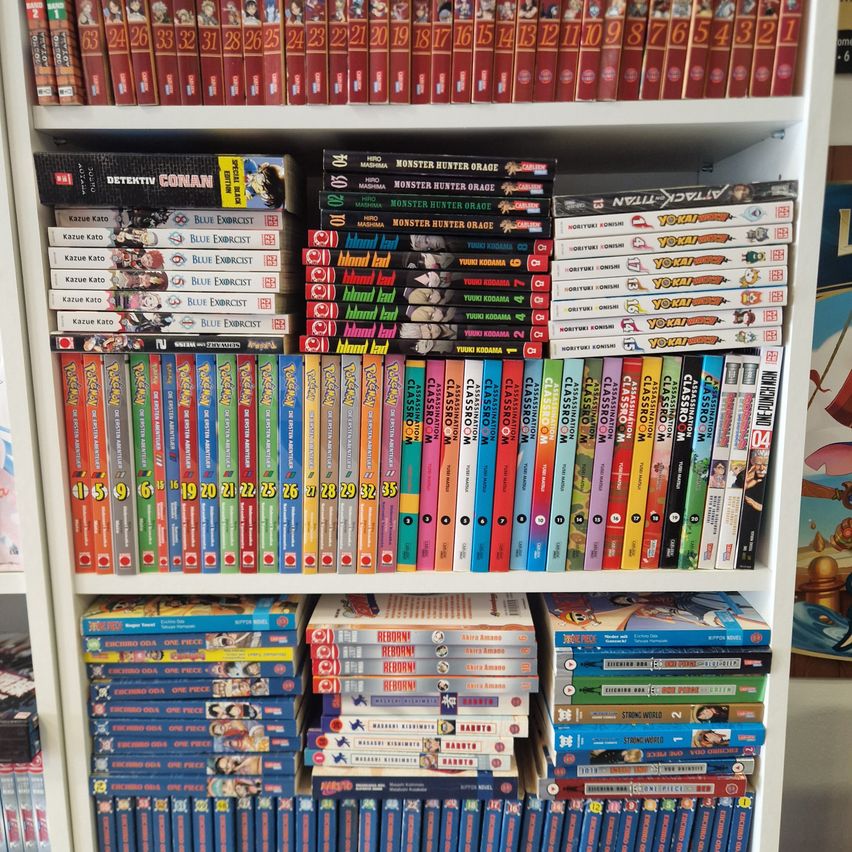 Ein weißer Bücherregal gefüllt mit Manga-Bänden, einschließlich Titeln wie 'Detective Conan' und 'Blue Exorcist', in Reihen angeordnet und aufeinander gestapelt.