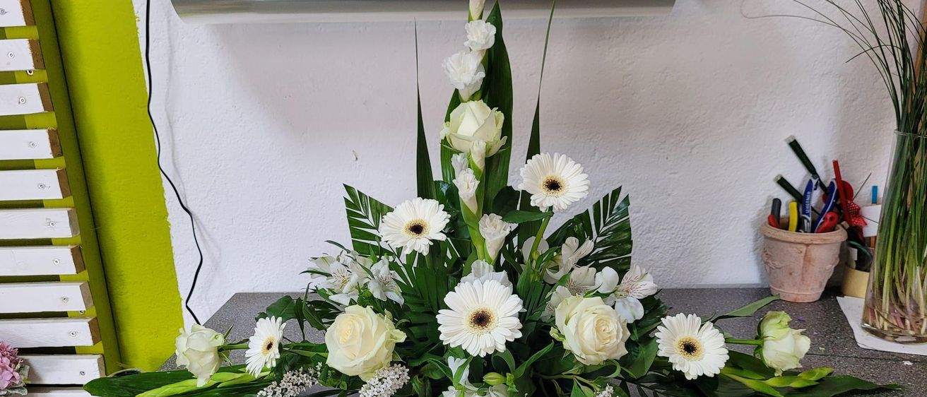 Eine große Blumenarrangement mit weißen Gänseblümchen und grünen Blättern auf einem Marmortisch, beleuchtet von einer silbernen Lampe oben.