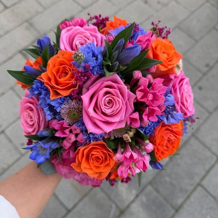 Ein farbenfroher Blumenstrauß mit rosa, orangen und blauen Blumen, gehalten von einer Hand auf einem Ziegelboden.
