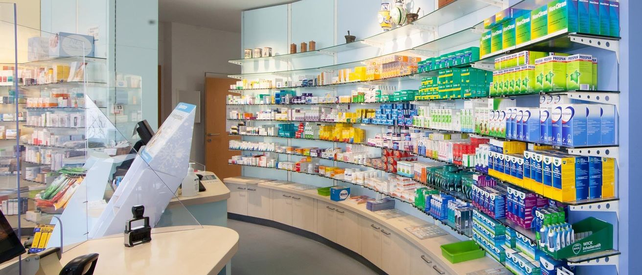 Eine Apotheke mit ordentlich angeordneten Regalen mit verschiedenen Medikamenten und Produkten, einer Holztür und einem Schreibtisch mit Computermonitor und Maus.