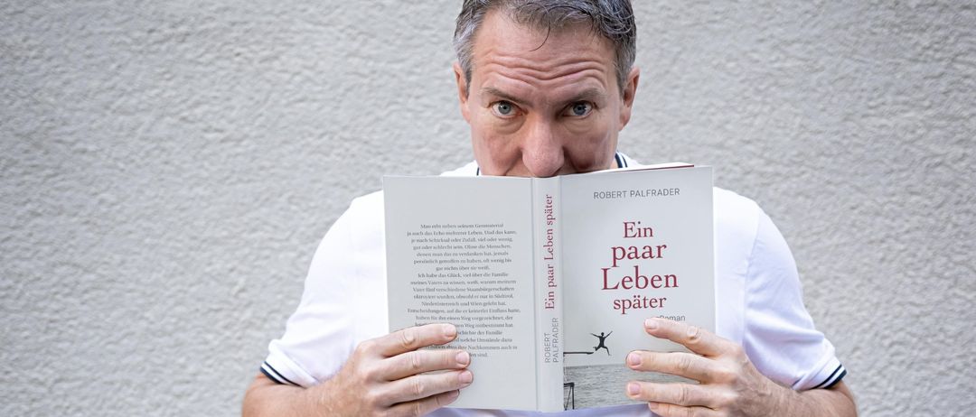 Ein Mann in einem weißen T-Shirt liest ein Buch mit dem Titel 'Ein paar Leben spater' von Robert Palfrader. Das Buchcover zeigt eine Person, die ins Wasser springt.