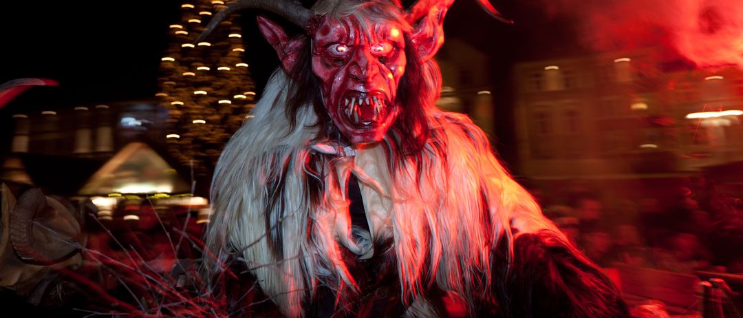 Eine Person in einer Dämonenmaske mit roten Augen und Hörnern steht vor einem beleuchteten Weihnachtsbaum in der Nacht.