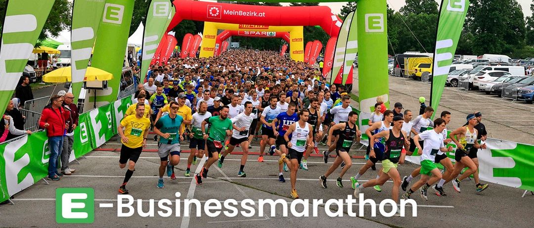 Läufer bei einem Marathon-Event, das unter einem Bogen mit 'Energie Steiermark' und 'Businessmarathon' geschrieben wird. Im Hintergrund sind viele Menschen und Autos zu sehen.