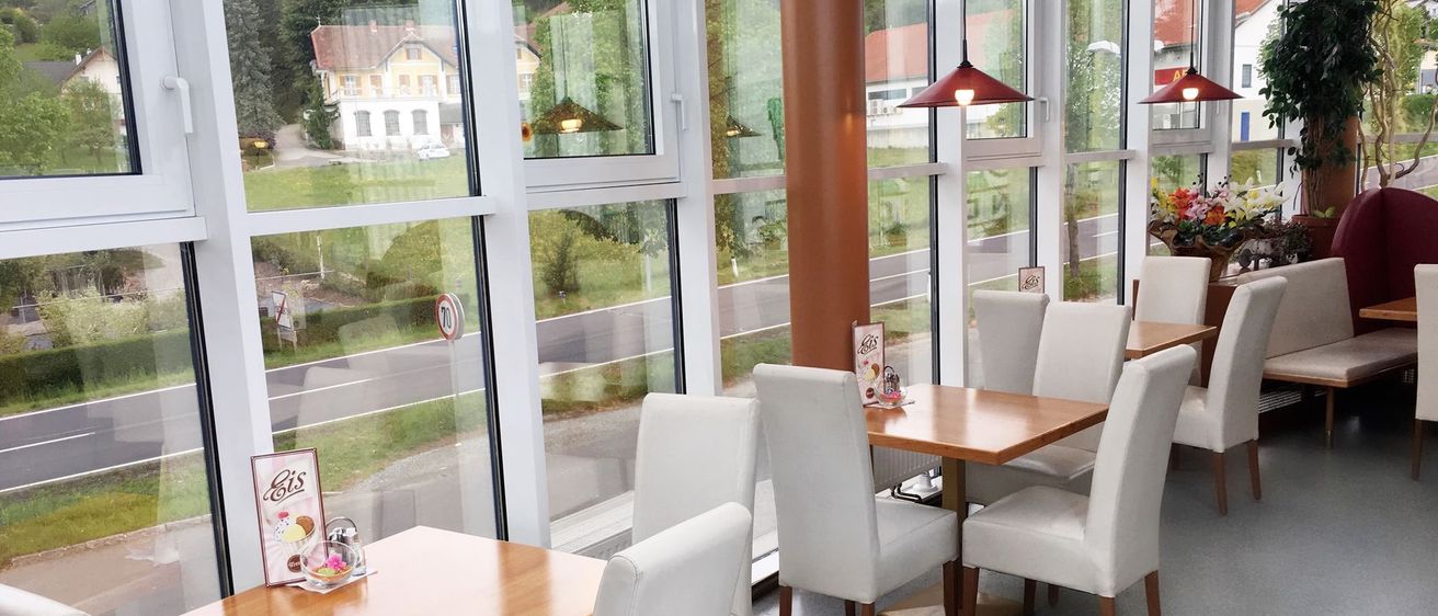 Ein Innenbereich zum Essen mit großen Fenstern, die auf eine Wiese und Berge blicken. Mehrere weiße Stühle und Tische sind angeordnet, mit dekorativen Lampen, die darüber hängen.