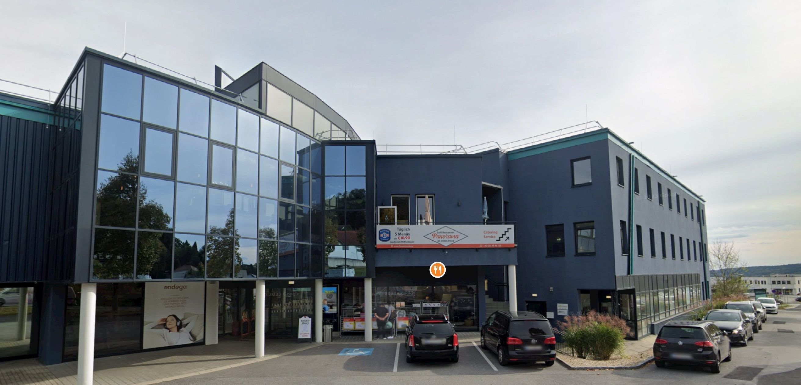 Ein modernes blaues Gebäude mit Glasfenstern und einem Schild, das Tagesmenüs €5,90 - €9,90 anzeigt. Es gibt einen Catering-Service und ein Restaurant namens Panorama. Vor dem Gebäude sind zwei Autos geparkt.