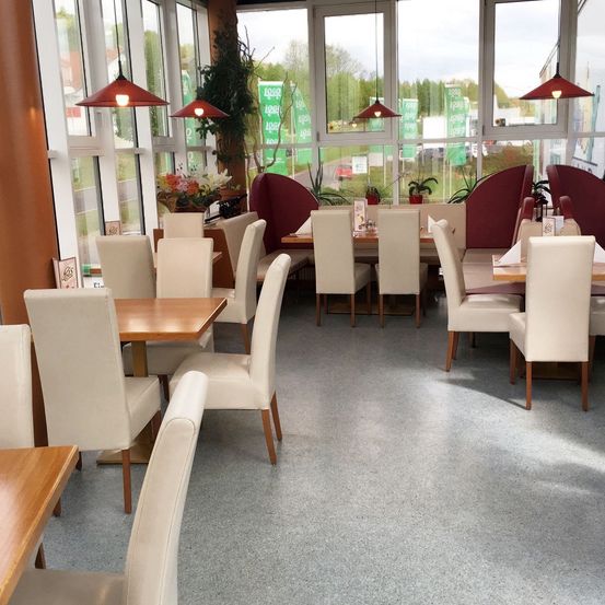 Ein Restaurant mit weißen Stühlen, Holztischen und großen Fenstern mit roten Pendelleuchten. Es gibt Pflanzen und Blumen, und der Boden ist grau.