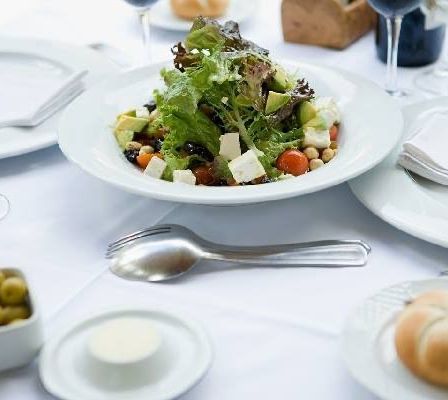 Ein Salat in einer weißen Schüssel mit Tomaten, Gurken, Feta und Oliven steht auf einem Tisch mit Tellern, Gläsern, einer Weinflasche und einem Brötchen.