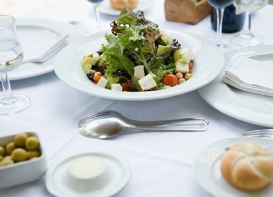 Ein Salat in einer weißen Schüssel mit Tomaten, Gurken, Feta und Oliven steht auf einem Tisch mit Tellern, Gläsern, einer Weinflasche und einem Brötchen.
