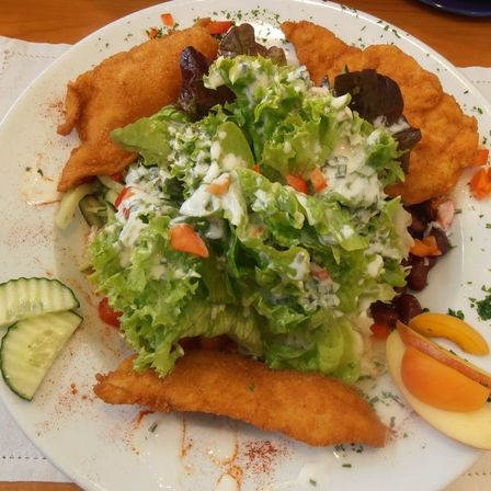 Ein weißer Teller mit einem gebratenen Hühnchengericht, Salat mit Dressing, Gurkenscheiben und Pfirsichscheiben auf einer Holzoberfläche.