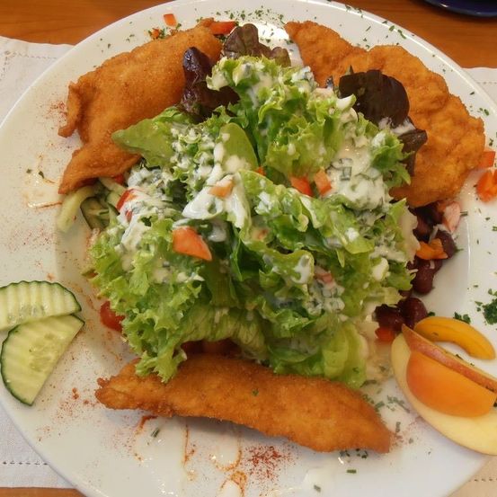 Ein weißer Teller mit einem gebratenen Hühnchengericht, Salat mit Dressing, Gurkenscheiben und Pfirsichscheiben auf einer Holzoberfläche.