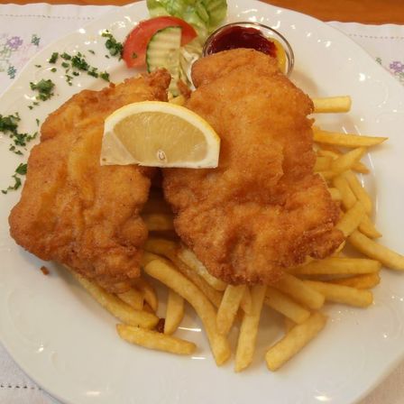 Ein Teller mit gebratenem Fisch, goldenen Pommes Frites und einer Zitronenscheibe obenauf. Ein kleiner Teller mit Sauce ist ebenfalls auf dem Teller.