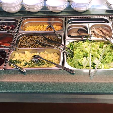 Ein Buffettisch mit verschiedenen Speisen. Die Schalen enthalten Mais, grüne Bohnen, Fleisch und Salat. Es sind Utensilien sichtbar.