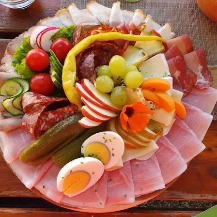 Ein Teller mit verschiedenen Lebensmitteln: geschnittener Schinken, Gurken, Trauben, Äpfel, Tomaten und Blumen. Das Essen steht auf einem Holztisch.