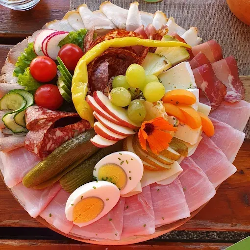 Ein Teller mit verschiedenen Lebensmitteln: geschnittener Schinken, Gurken, Trauben, Äpfel, Tomaten und Blumen. Das Essen steht auf einem Holztisch.